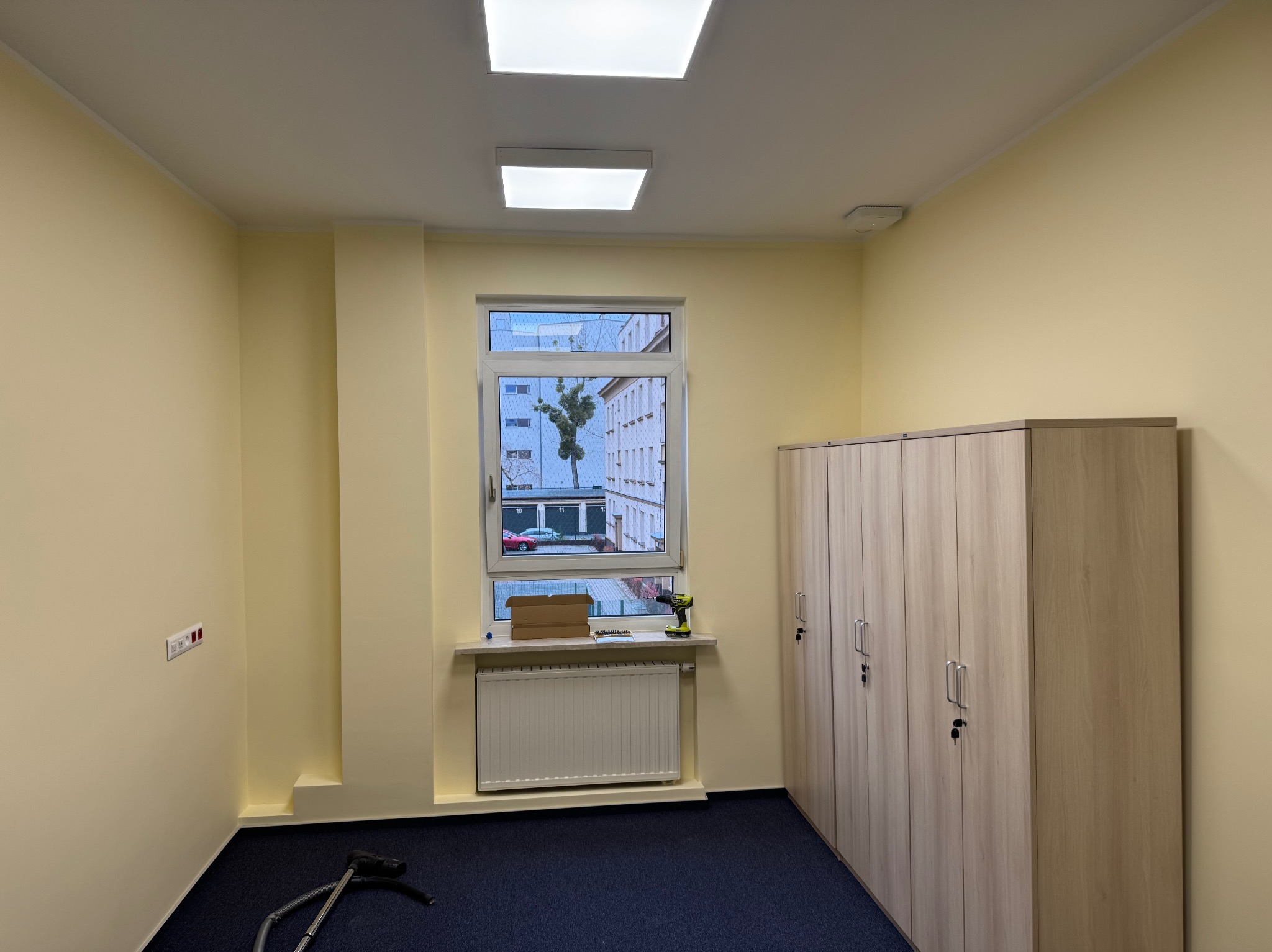 Pomieszczenie po malowaniu ścian na jasnożółty kolor. Widoczne szafy, okno, grzejnik, narzędzia i odkurzacz. Na ścianie gniazdka elektryczne.