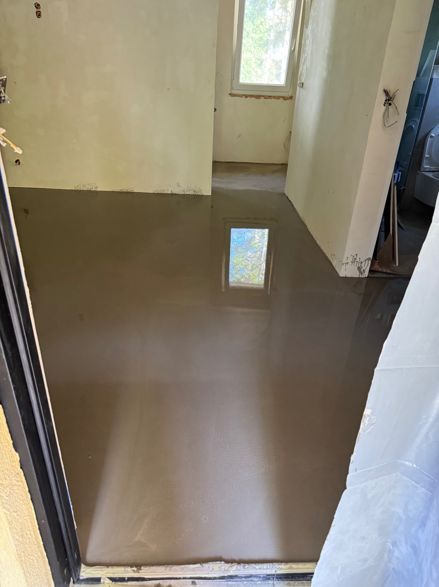 Wylana, mokra wylewka samopoziomująca w pomieszczeniu z oknem. Widoczne ślady po zabrudzeniach na ścianach. Część okna odbija się w mokrej powierzchni.