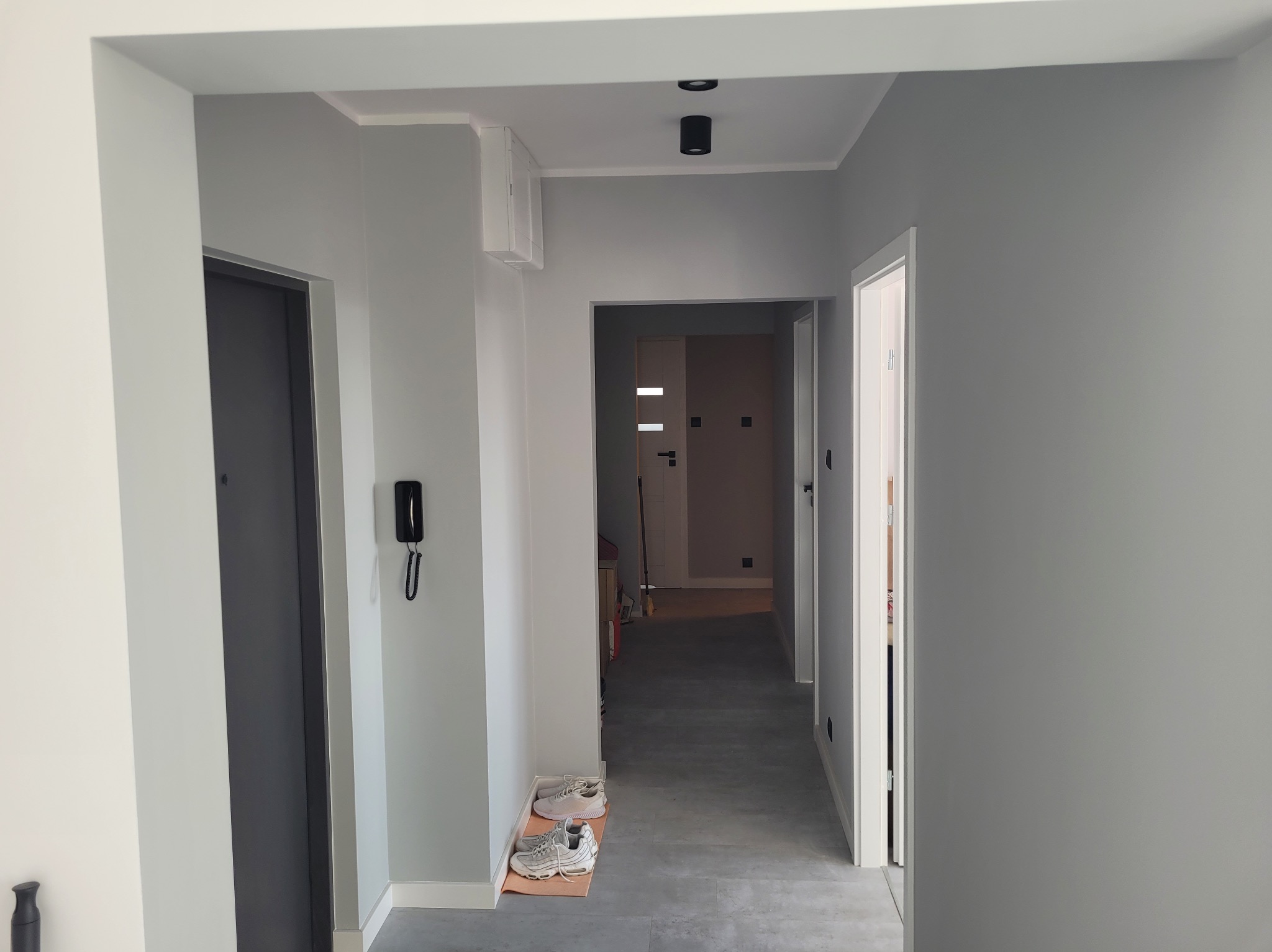 Szary korytarz po remoncie z drzwiami, oświetleniem sufitowym i parą butów na wycieraczce. Świeżo pomalowane ściany i minimalistyczny design.