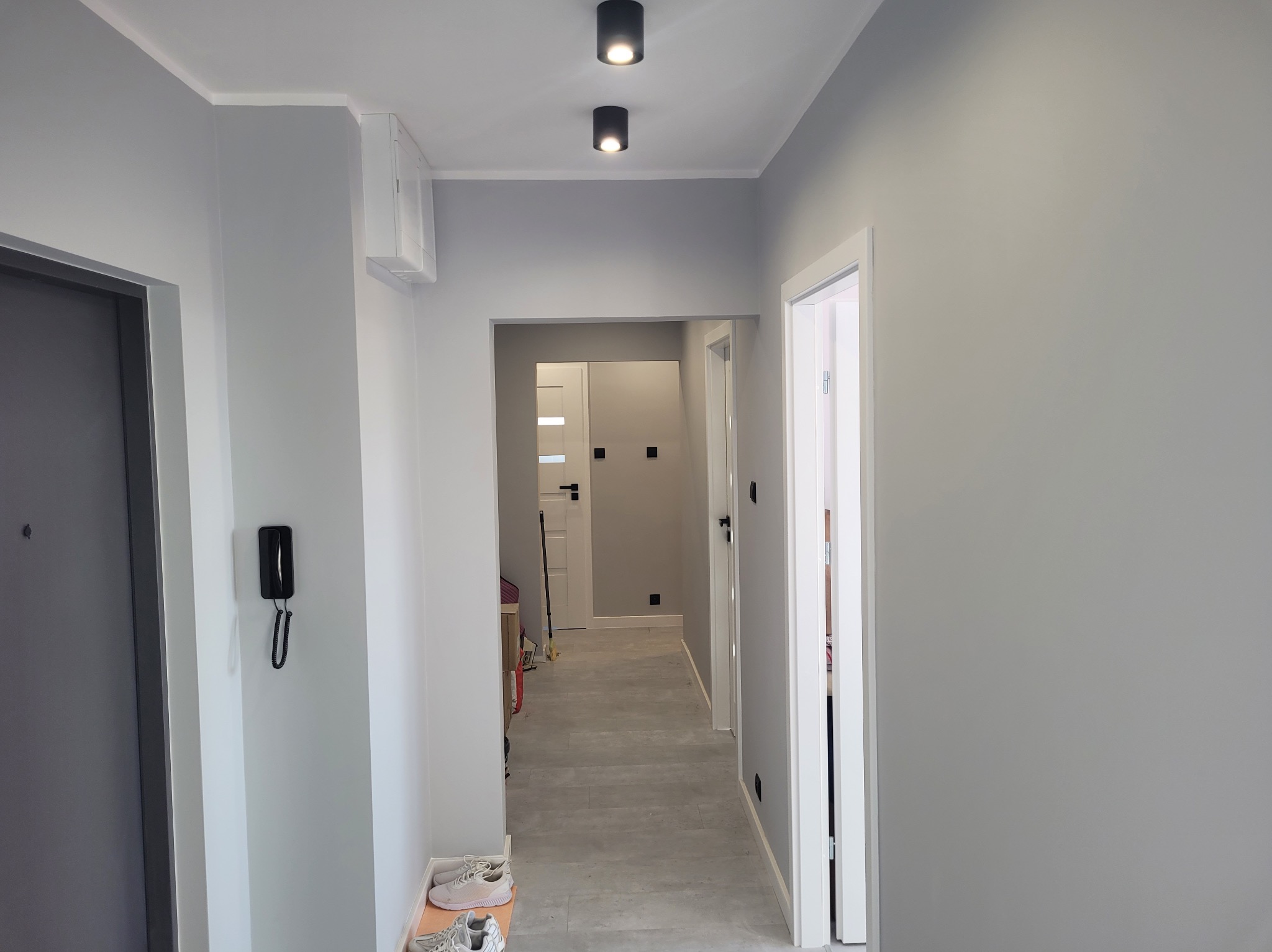 Nowoczesny, szary przedpokój po remoncie: gładkie ściany, podwieszany sufit z oświetleniem punktowym, minimalistyczny design z interkomem przy wejściu.