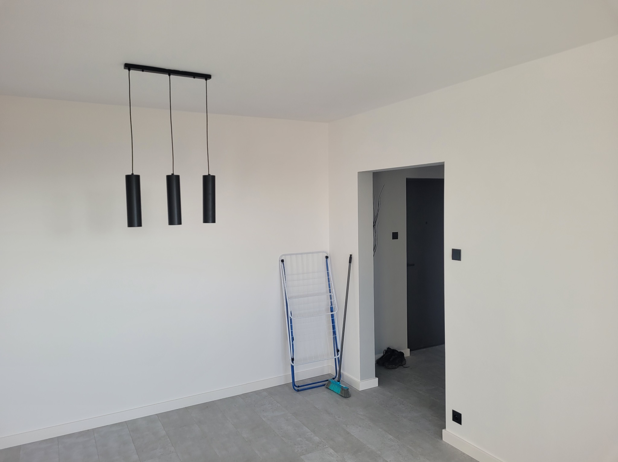 Wnętrze po remoncie: białe ściany, szara podłoga, minimalistyczna lampa. Widoczny wieszak na pranie i drzwi. Czyste, odświeżone pomieszczenie.