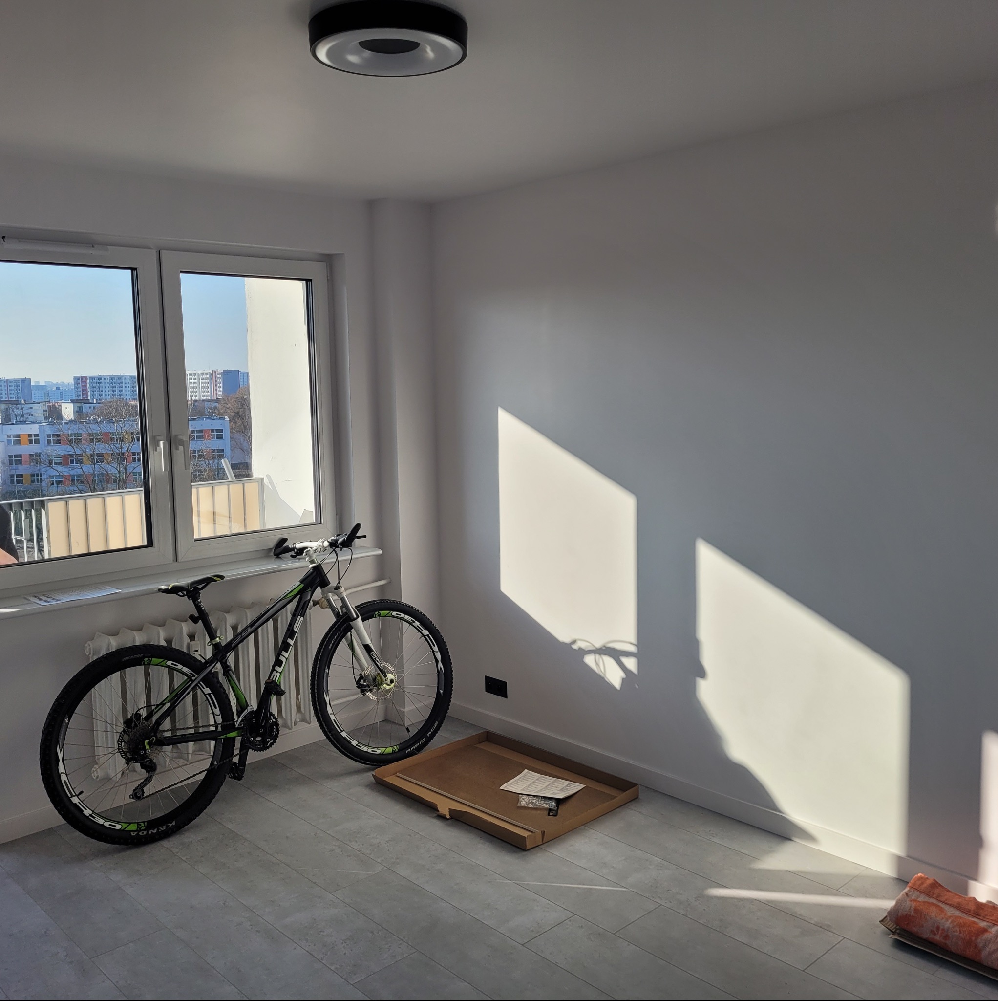 Jasne wnętrze pokoju po remoncie: białe ściany, okno z widokiem na miasto, rower oparty o ścianę, karton z elementami montażowymi na podłodze. Minimalizm i czystość.