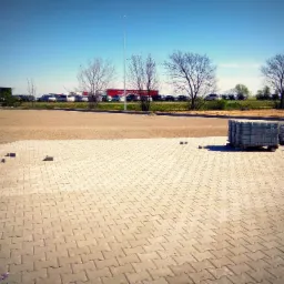 Układanie szarej kostki brukowej na parkingu, palety z kostką w tle, widoczny fragment podbudowy, drzewa bez liści i zaparkowane samochody.