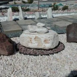 Kompozycja z kamieni o różnych fakturach i kolorach na tle wysypanego żwiru, z elementami ścieżki z płyt w tle.