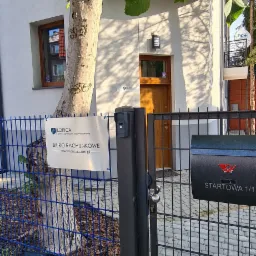 Tablica informacyjna Biura Rachunkowego 'Lorica' przy wejściu do budynku, widoczna przez niebieskie ogrodzenie i czarną skrzynkę na listy.