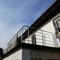 Balkon z nierdzewną, poziomą balustradą, widok z dołu na błękitne niebo, elewacja budynku w trakcie remontu, widoczne ocieplenie i fragmenty starego muru.