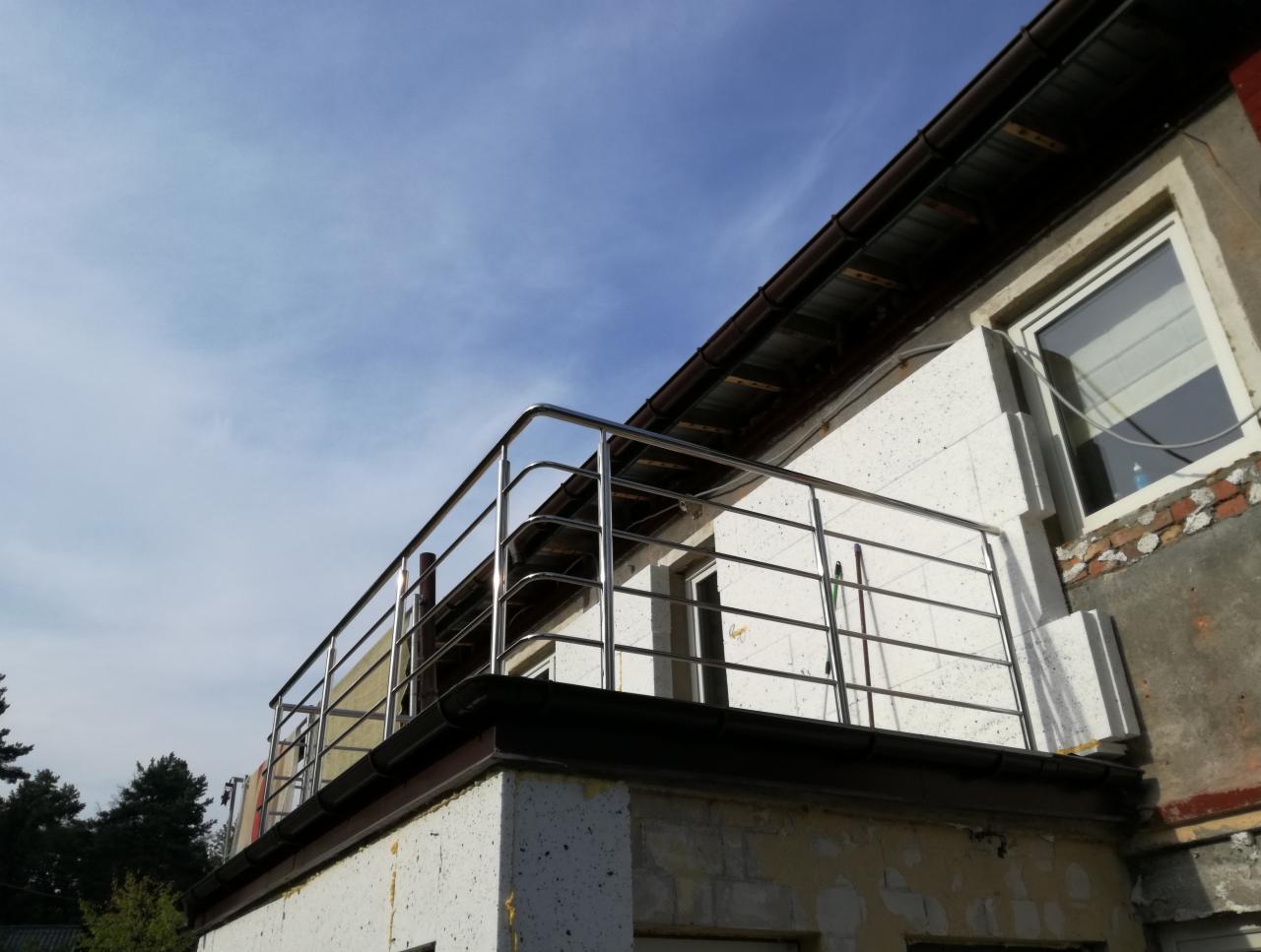 Balkon z nierdzewną, poziomą balustradą, widok z dołu na błękitne niebo, elewacja budynku w trakcie remontu, widoczne ocieplenie i fragmenty starego muru.