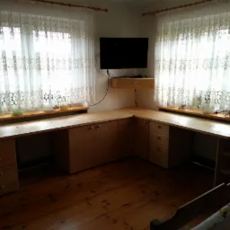 Jasny, drewniany narożny biurko z szafkami i szufladami, umieszczone pod dwoma oknami z koronkowymi firankami, z telewizorem na ścianie i drewnianą podłogą.