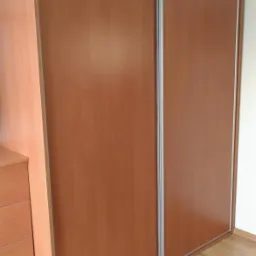 Drewniana szafa przesuwna z dwoma drzwiami i srebrnymi prowadnicami, umieszczona w rogu pokoju, widoczna z boku, z fragmentem komody po lewej stronie i łóżkiem w dolnej części kadru.