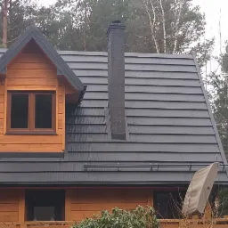 Dom z drewnianą elewacją i dachem pokrytym szarą dachówką karpiówką oraz czarnym kominem z cegły, widok na las w tle.