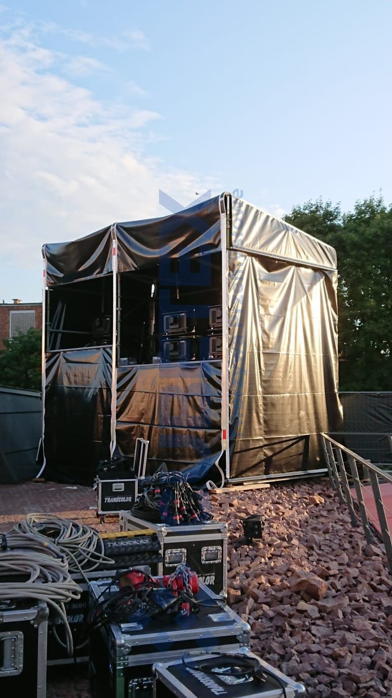 Tymczasowa scena z czarną plandeką, widoczne skrzynie transportowe z kablami i sprzętem oświetleniowym, na tle nieba i drzew.