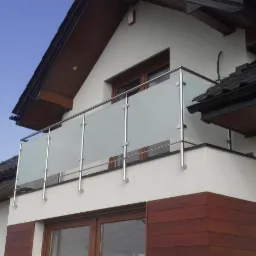 Balkon z matową szklaną balustradą i nierdzewnymi słupkami, widok z dołu na tle jasnego błękitnego nieba i fragmentu dachu domu.