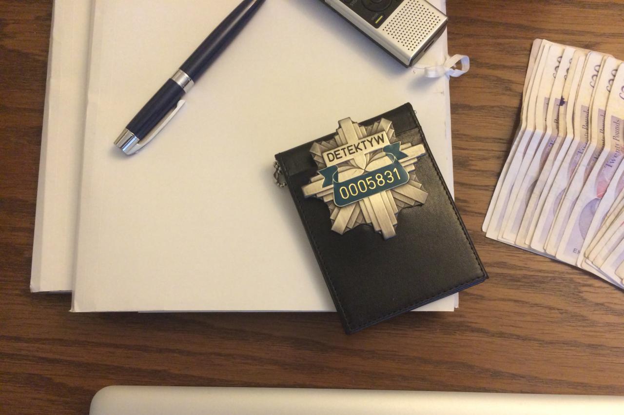 Legitymacja detektywa z numerem 0005831 leżąca na czarnym skórzanym etui, obok pliku dokumentów, długopisu, dyktafonu i pliku banknotów dwudziestofuntowych na drewnianym biurku.