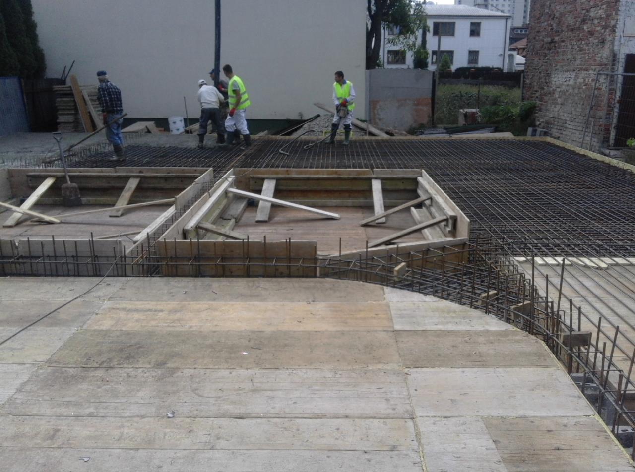 Przygotowanie szalunków i zbrojenia na budowie, widoczni robotnicy w kamizelkach odblaskowych, w tle budynki mieszkalne.