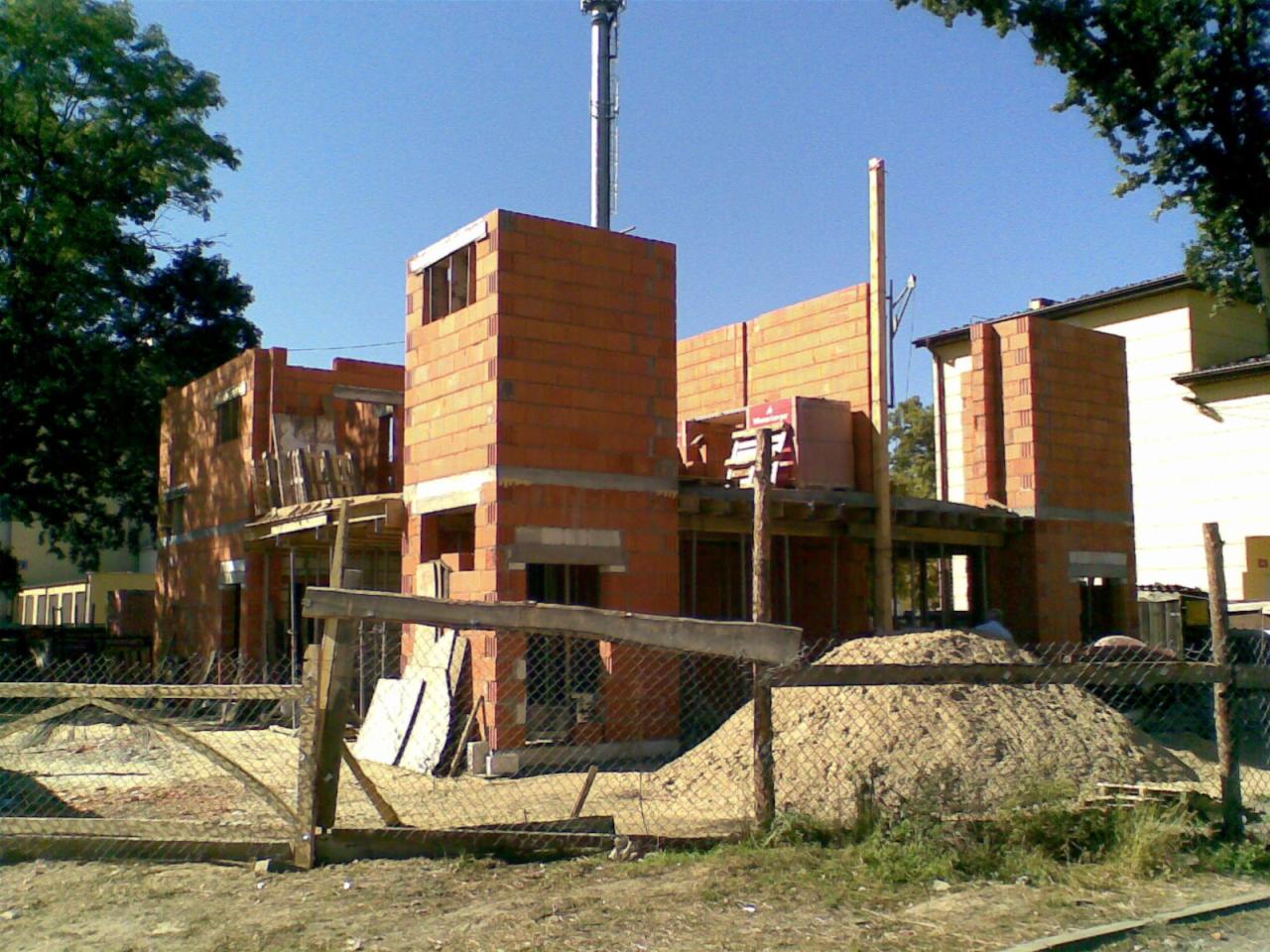 Wczesny etap budowy domu z cegły ceramicznej, widoczne ściany nośne i kominy, zabezpieczony teren budowy z hałdą piasku.