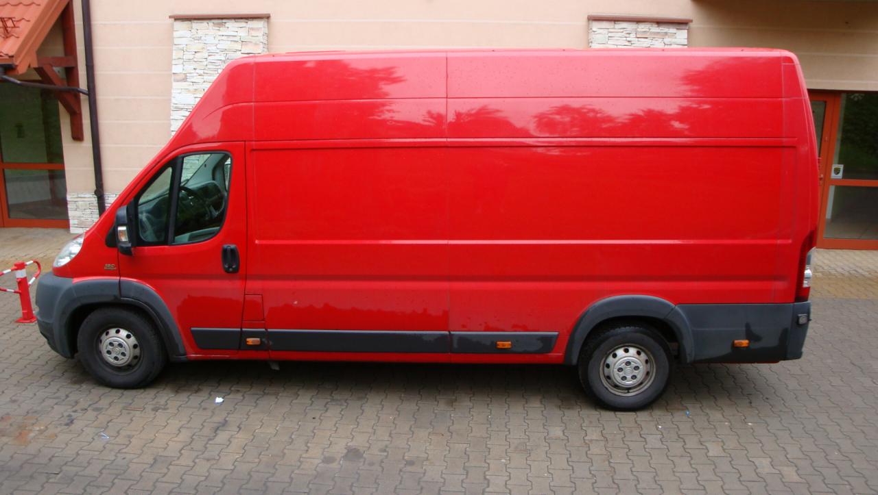 Czerwony samochód dostawczy zaparkowany na brukowanym podjeździe przed budynkiem z beżową elewacją i dwoma oknami z kamienną obudową.