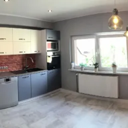 Nowoczesna kuchnia z szarymi szafkami dolnymi, białymi górnymi, ceglaną ścianą i drewnianą podłogą, oświetlona wiszącymi lampami z żarówkami Edisona.