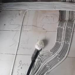 Białe kable elektryczne w peszlach ułożone równolegle wzdłuż ściany z widocznymi oznaczeniami ołówkiem, czarne kable wychodzące ze ściany zabezpieczone pianką montażową.