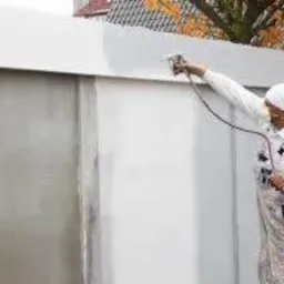 Pracownik w kombinezonie ochronnym i chuście na głowie, aplikuje farbę natryskowo na zewnętrzną ścianę budynku. Widoczny pistolet natryskowy i przewód doprowadzający farbę. Częściowo pomalowana...