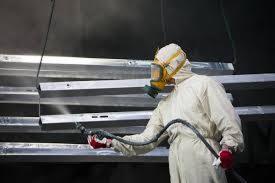 Pracownik w kombinezonie ochronnym i masce lakierniczej natryskuje farbę na metalowe profile zawieszone w warsztacie, używając pistoletu lakierniczego z długim wężem.