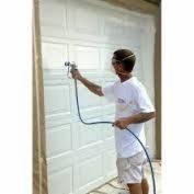 Pracownik w Toruniu, w białej koszulce i okularach ochronnych, natryskowo aplikuje białą farbę na segmentową bramę garażową, zabezpieczoną folią malarską.