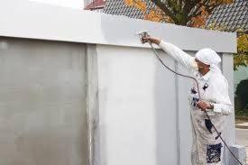 Pracownik w kombinezonie ochronnym i chuście na głowie, aplikuje farbę natryskowo na zewnętrzną ścianę budynku. Widoczny pistolet natryskowy i przewód doprowadzający farbę. Częściowo pomalowana...