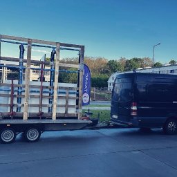 Wierzbicki Transport InstalLoft - Czarny van z przyczepą przewożącą białe płyty zabezpieczone drewnianą konstrukcją, flaga firmy widoczna w tle.
