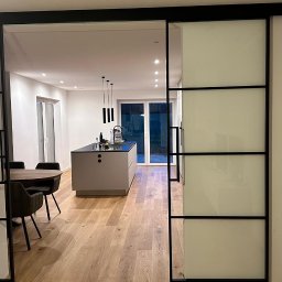 Wierzbicki Transport InstalLoft - Nowoczesne przesuwne drzwi wewnętrzne z matowego szkła i czarnych profili, oddzielające jadalnię od kuchni z wyspą, widok na starannie wykończone wnętrze.