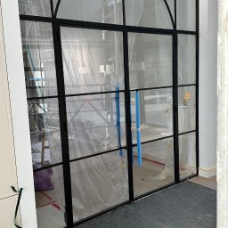 Wierzbicki Transport InstalLoft - Dwuskrzydłowe drzwi wewnętrzne z czarnymi szprosami i łukowym nadświetlem, zabezpieczone folią ochronną podczas remontu, widoczny dywanik i fragment rusztowania w tle.