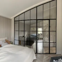 Duże, przeszklone drzwi wewnętrzne z czarnymi szprosami, oddzielające sypialnię od schodów, z widocznym łóżkiem z pluszowym leniwcem i parkietem w jodełkę.