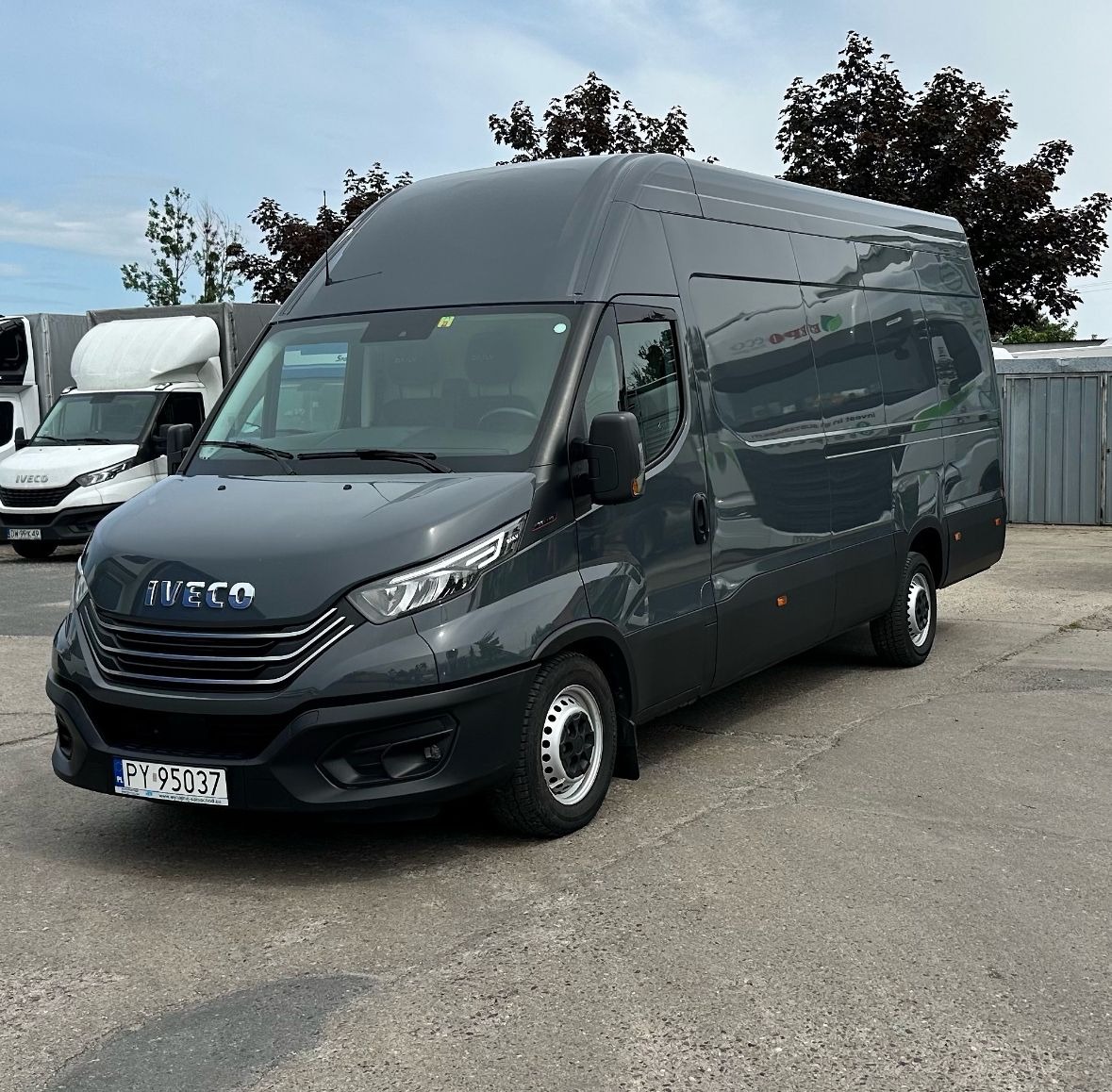 Szary samochód dostawczy Iveco z wysokim dachem zaparkowany na betonowym placu, z widocznymi innymi pojazdami w tle i drzewami w tle.