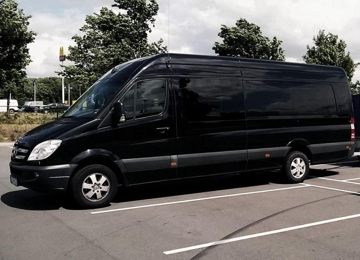 Czarny, lśniący bus Mercedes-Benz Sprinter zaparkowany na parkingu, z widocznym znakiem McDonald's w tle.