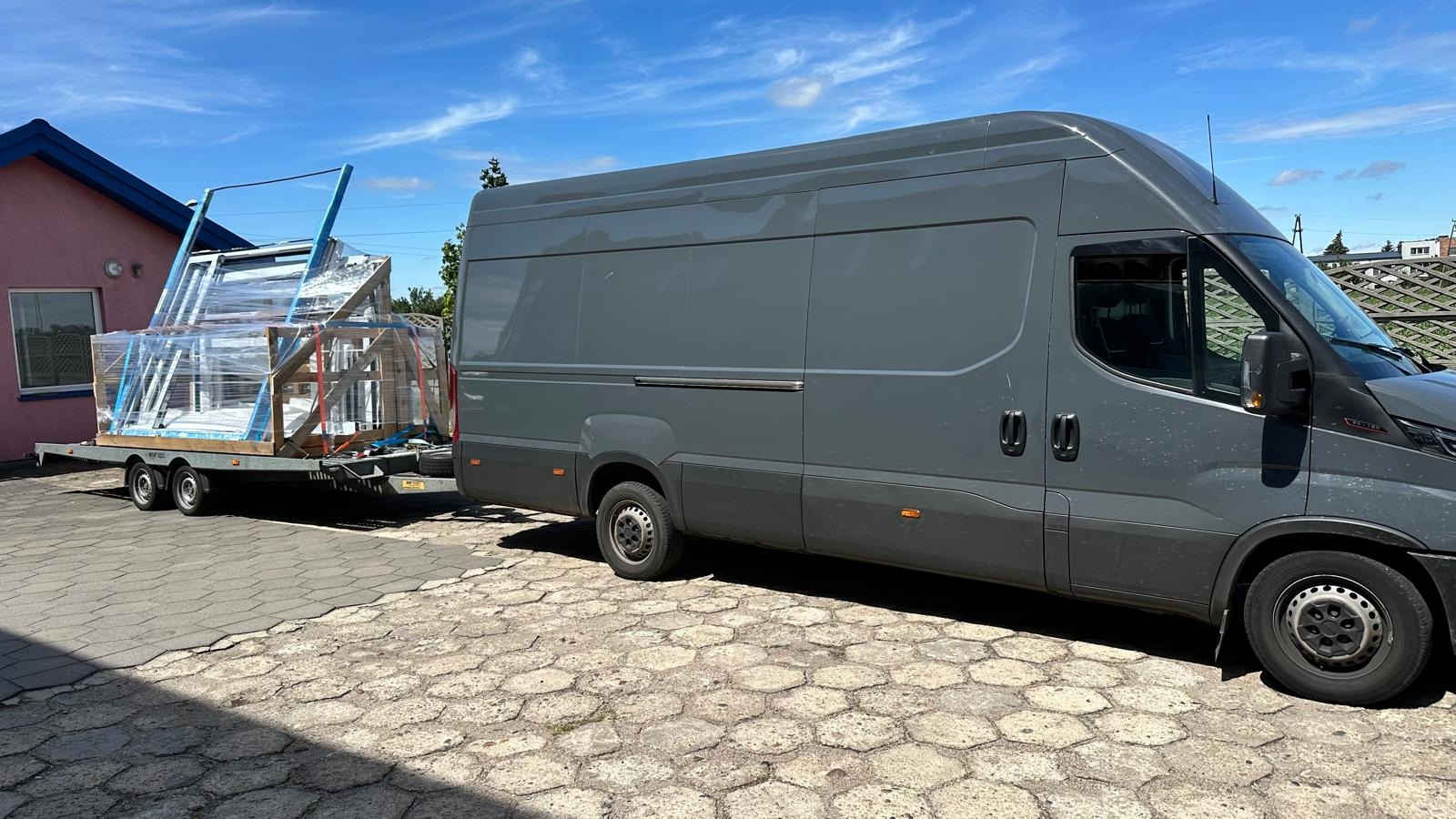 Szary bus z przyczepą przewożącą okna, zabezpieczone folią, na tle domu i błękitnego nieba.