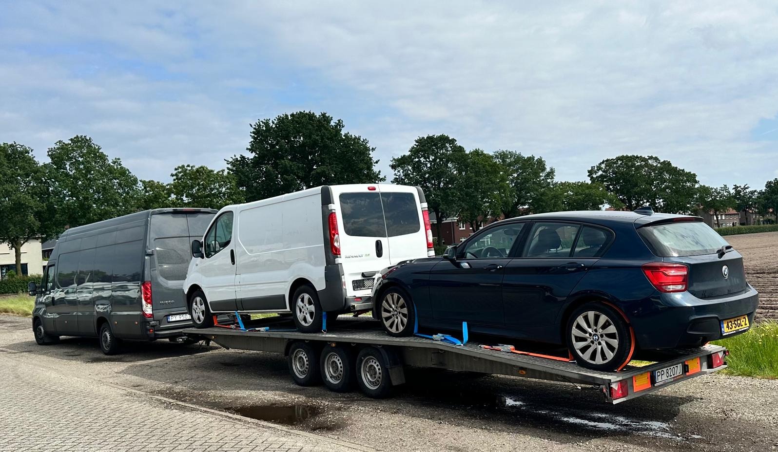 Laweta z dwoma busami i samochodem osobowym, zabezpieczonymi pasami transportowymi, na tle drzew i pola.