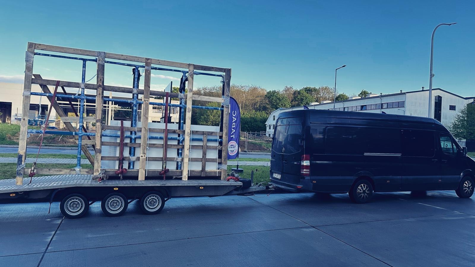 Czarny van z przyczepą przewożącą białe płyty zabezpieczone drewnianą konstrukcją, flaga firmy widoczna w tle.