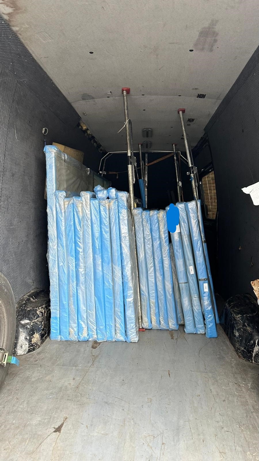 Wnętrze busa wypełnione po brzegi spakowanymi, zabezpieczonymi folią elementami, prawdopodobnie drzwiami, widoczne metalowe podpory sufitowe, opona i brudna podłoga.