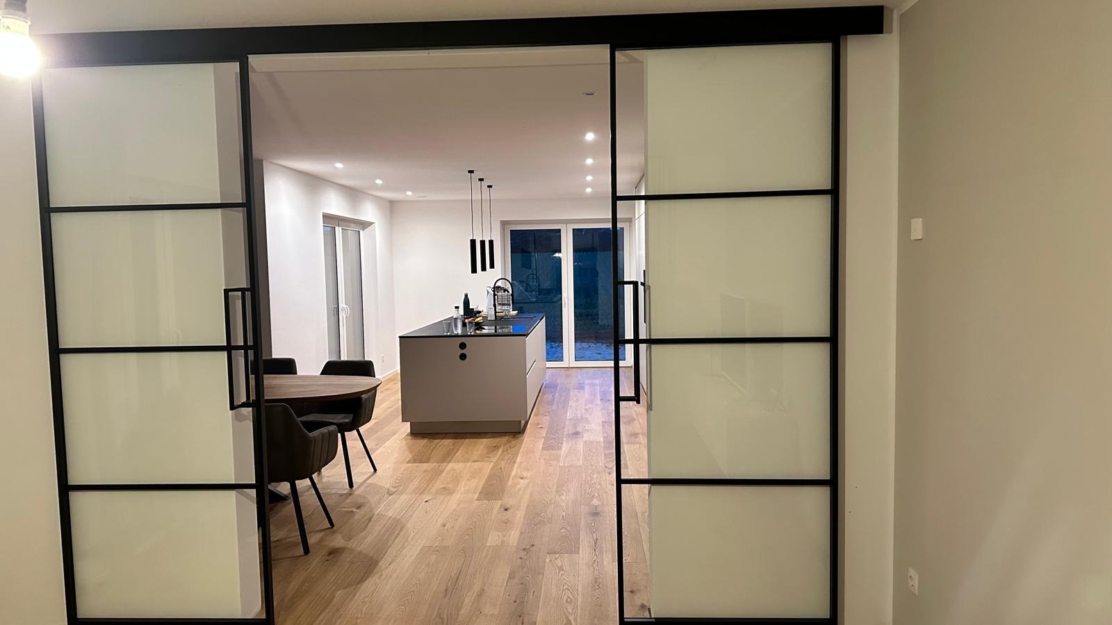 Nowoczesne przesuwne drzwi wewnętrzne z matowego szkła i czarnych profili, oddzielające jadalnię od kuchni z wyspą, widok na starannie wykończone wnętrze.