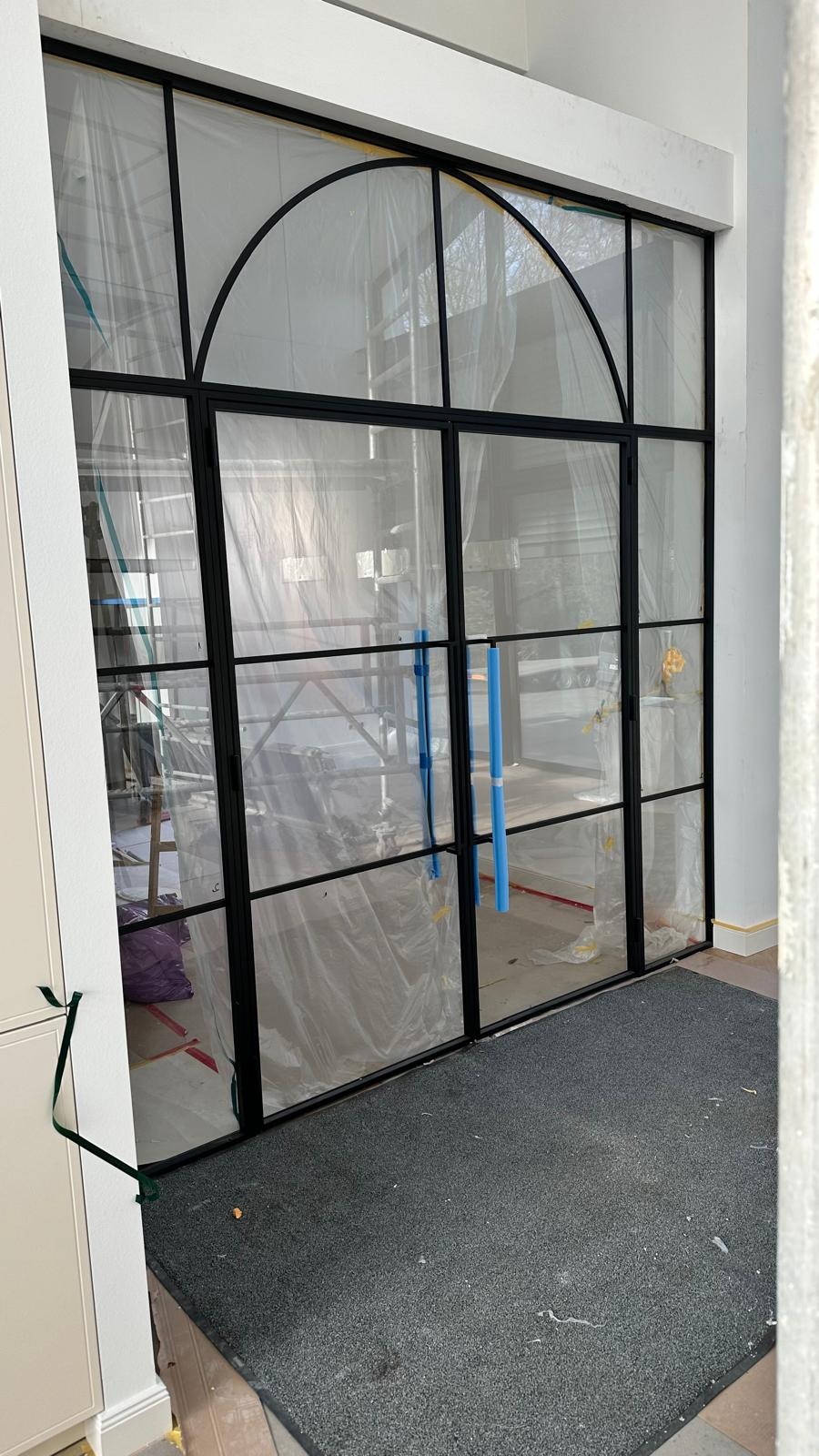 Dwuskrzydłowe drzwi wewnętrzne z czarnymi szprosami i łukowym nadświetlem, zabezpieczone folią ochronną podczas remontu, widoczny dywanik i fragment rusztowania w tle.