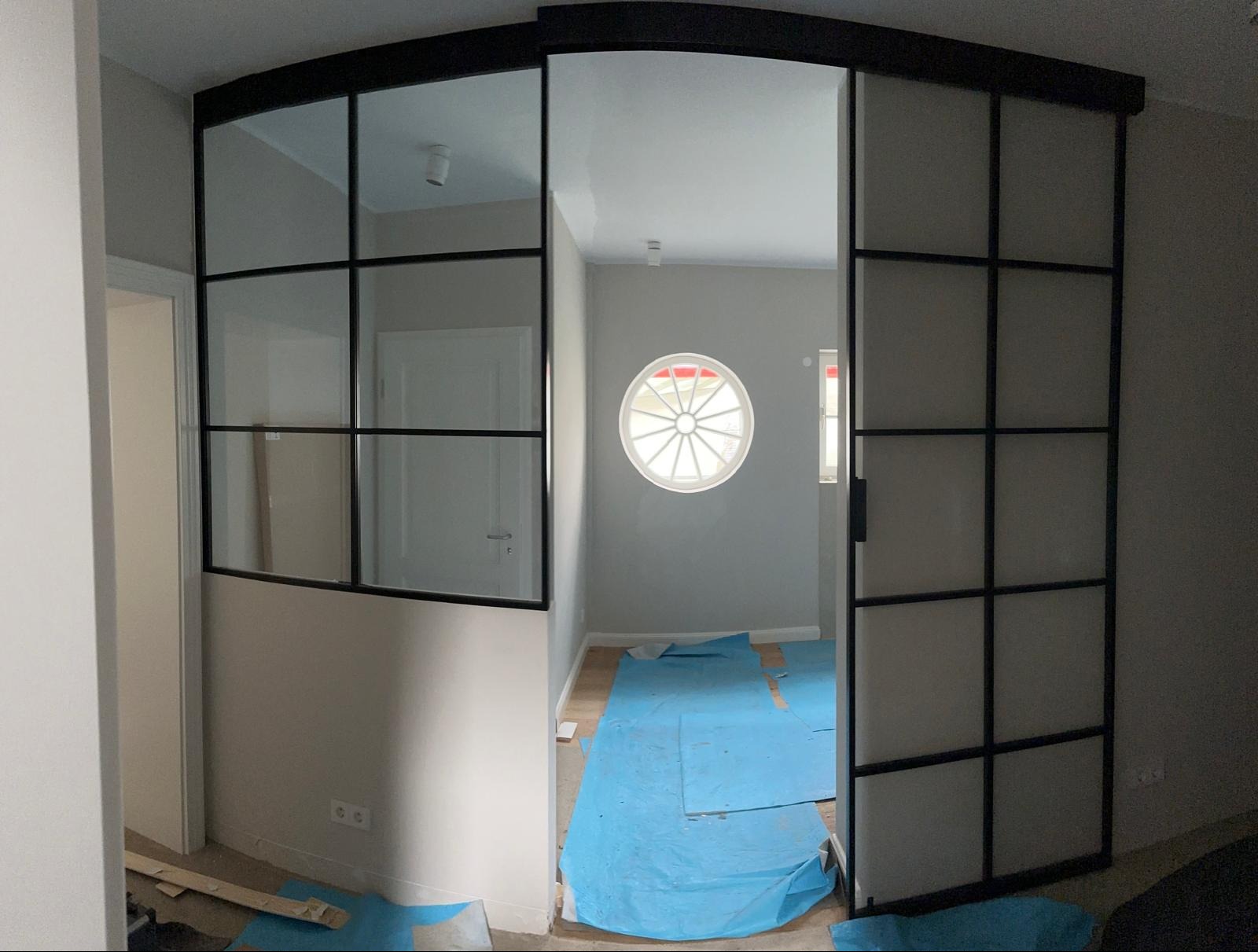 Przesuwne drzwi wewnętrzne w stylu industrialnym z czarnymi szprosami, częściowo zasłonięte folią ochronną na podłodze, widoczne okrągłe okno w tle.