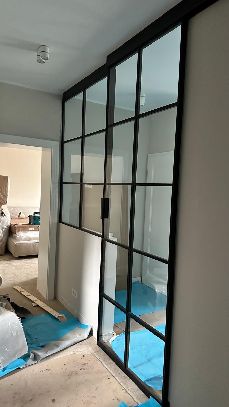 Nowoczesne przesuwne drzwi wewnętrzne z czarnymi szprosami i szklanymi panelami, widoczne podczas montażu, z zabezpieczoną podłogą i częściowo spakowanymi meblami w tle.
