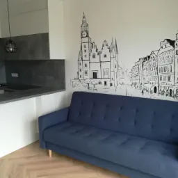 Wnętrze mieszkania z aneksem kuchennym i niebieską sofą na tle ściany z czarno-białym rysunkiem panoramy miasta.