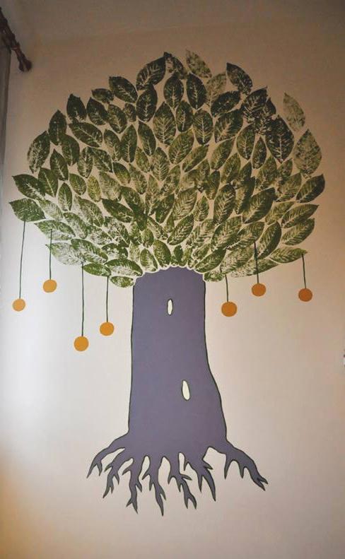 Malowidło na ścianie przedstawiające drzewo z zielonymi liśćmi odbitymi stemplem i fioletowym pniem, z pomarańczowymi owocami wiszącymi na cienkich sznurkach.