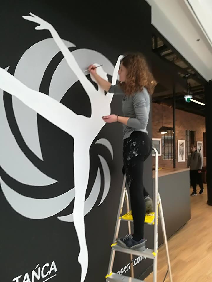 Kobieta na drabinie maluje pędzlem duży, biały, stylizowany wizerunek tancerki na czarnej ścianie wewnątrz budynku. Częściowo widoczne logo firmy 'TANCA' w lewym dolnym rogu.