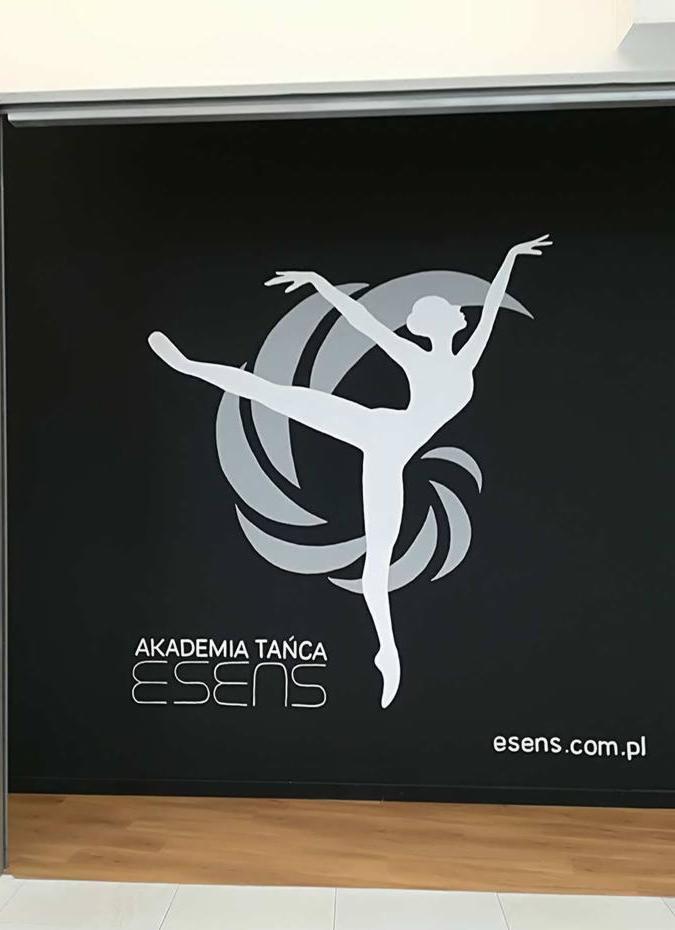 Naklejka ścienna z sylwetką tancerki w baletowej pozie, logo Akademii Tańca Esens na czarnym tle w sali tanecznej.