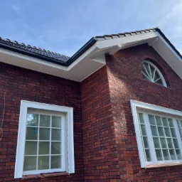 Fragment domu z czerwonej cegły z białymi oknami, czarnym dachem i rynnami, oraz półokrągłym oknem na poddaszu.