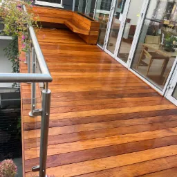 Błyszczący, świeżo zaimpregnowany taras z egzotycznego drewna z balustradą ze stali nierdzewnej, z widocznym siedziskiem/skrzynią na rośliny z tego samego drewna, z kwiatami w donicach...