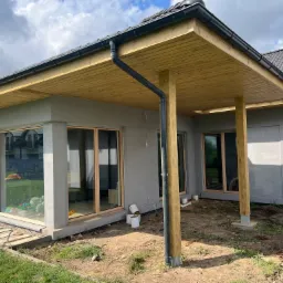 Zadaszony taras przy nowoczesnym domu z szarą elewacją, widoczne drewniane podpory i podbitka, czarne rynny, teren wokół w trakcie zagospodarowywania, widoczne puszki po farbie.