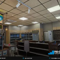 Wnętrze lokalu handlowego z widocznymi systemami wentylacji podwieszonymi pod sufitem, oświetleniem punktowym i kratką wentylacyjną, z pustymi regałami chłodniczymi na piwo i napoje.