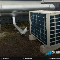 Zewnętrzna jednostka klimatyzacji zamontowana na betonowych podstawach, z widocznymi przewodami i elementami instalacji na dachu budynku komercyjnego.