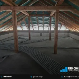 Poddasze w trakcie instalacji systemu wentylacyjnego: widoczne rury wentylacyjne ułożone na podłodze, drewniana konstrukcja dachu i ściany z bloczków. Układ rur sugeruje rozprowadzenie powietrza po...